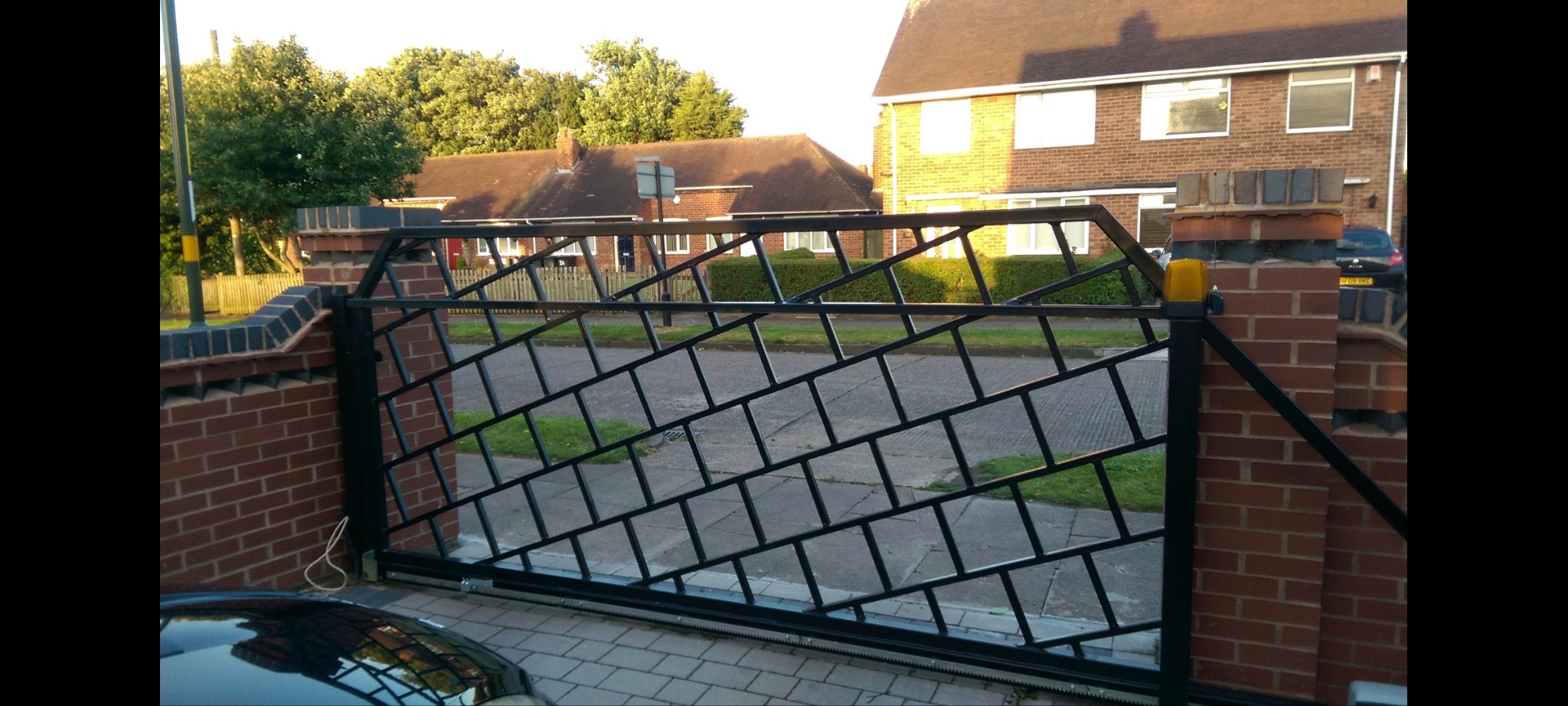 Czarna brama wjazdowa przesuwna z metalową kratką o ukośnym wzorze, zamontowana na ceglanych słupkach w Kotach, widok z zewnątrz posesji.