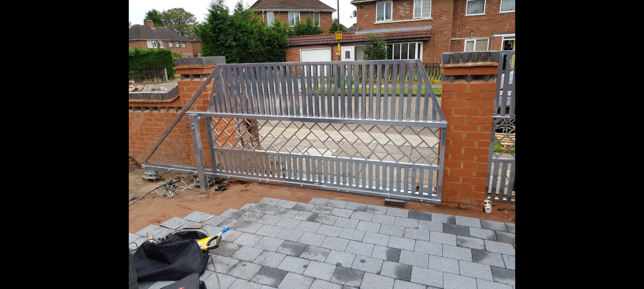 Surowa, ocynkowana brama posesyjna z metalowymi prętami i kratą w romby, w trakcie montażu, widoczne ceglane słupki i ułożona kostka brukowa.