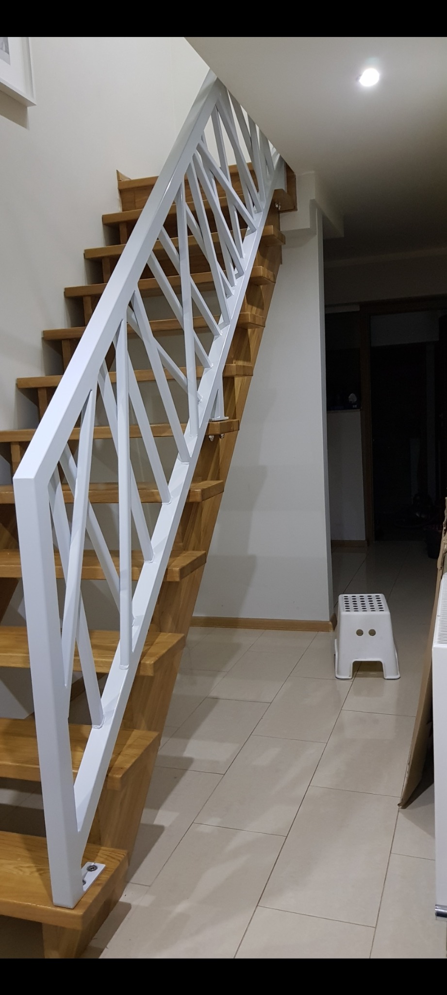 Drewniane schody z białą, geometryczną balustradą wewnątrz domu, widoczne z boku, z fragmentem białego taboretu w tle.