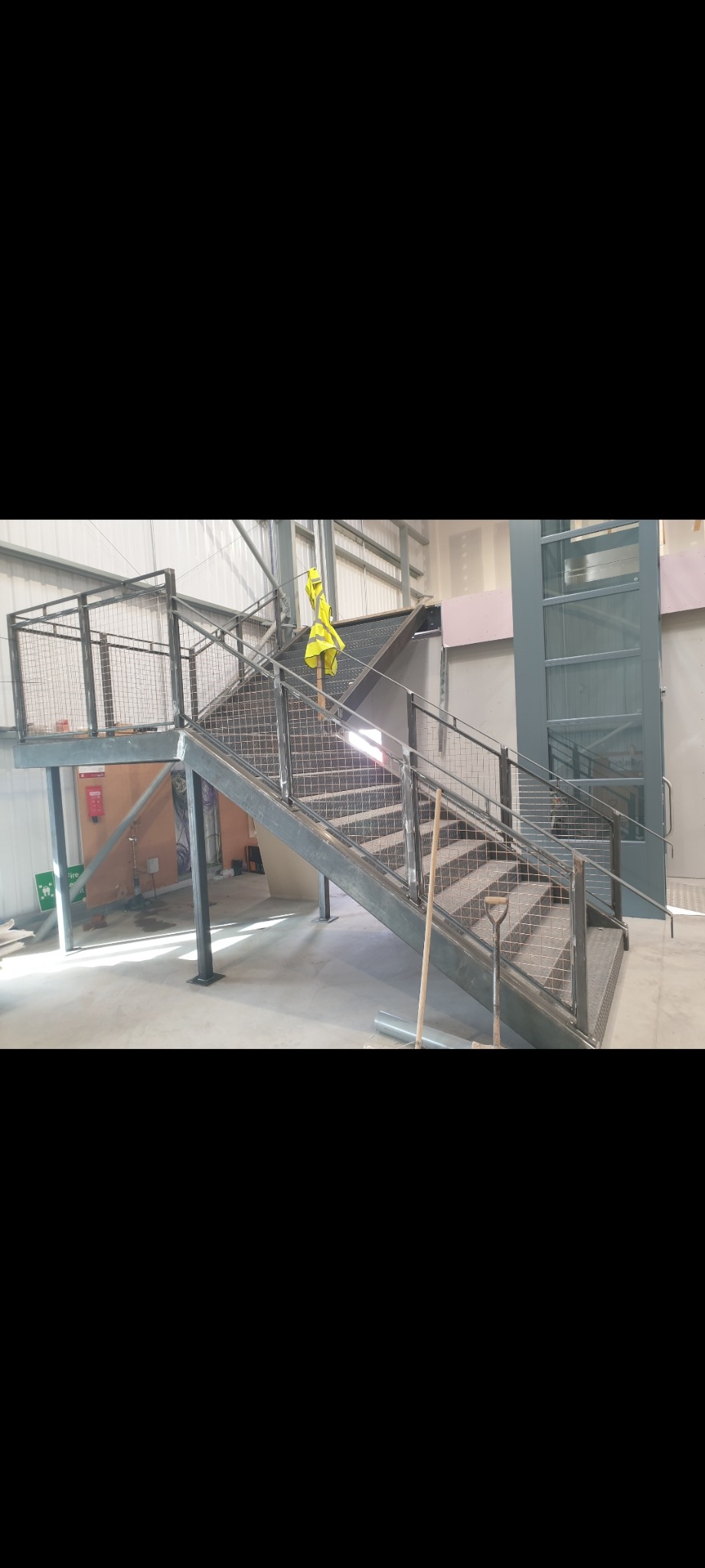Metalowe schody z siatkową balustradą w surowym wnętrzu hali, z żółtą kamizelką odblaskową wiszącą na górnym poziomie, widoczne narzędzia budowlane.