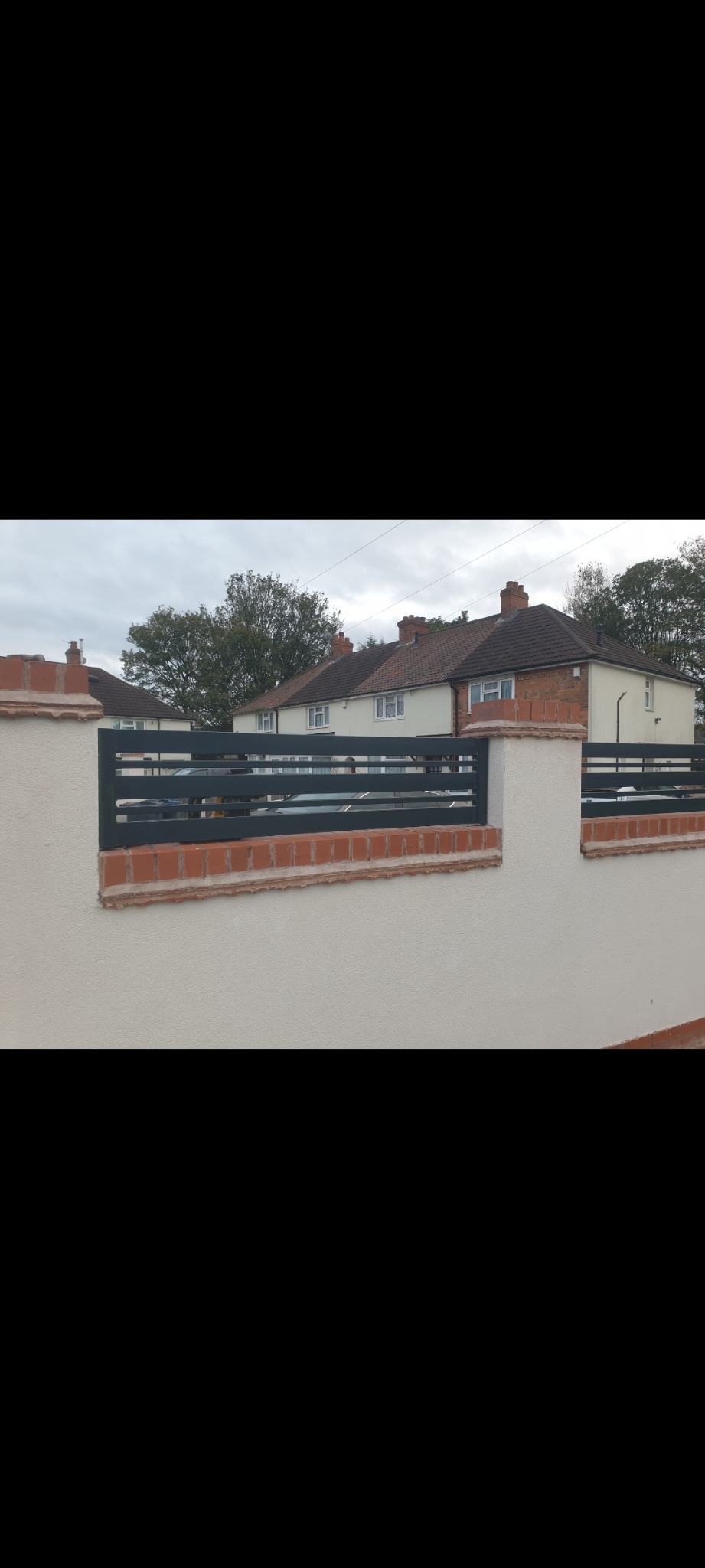 Nowoczesne ogrodzenie z poziomych, grafitowych elementów metalowych, osadzone na słupkach z białego tynku i cegły klinkierowej, na tle domów jednorodzinnych.