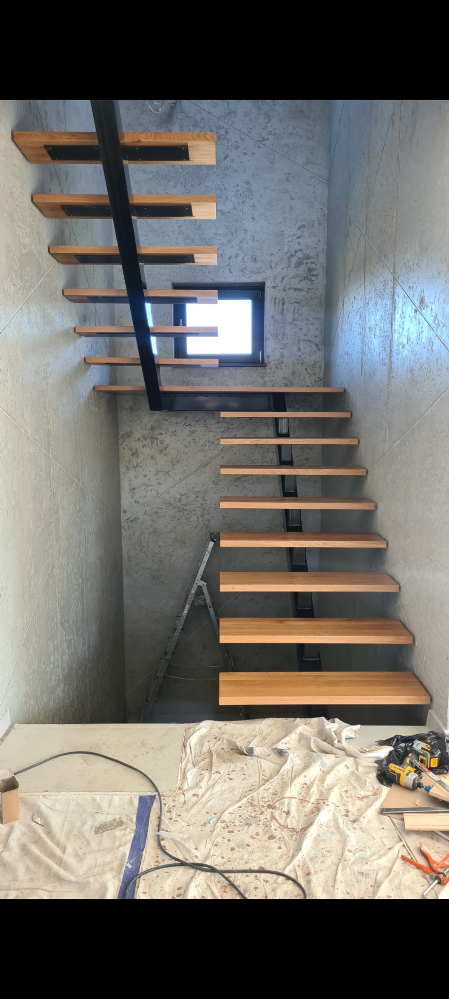 Schody wiszące z drewnianymi stopniami i czarną metalową konstrukcją, widoczne narzędzia i drabina w trakcie montażu na tle surowych betonowych ścian.