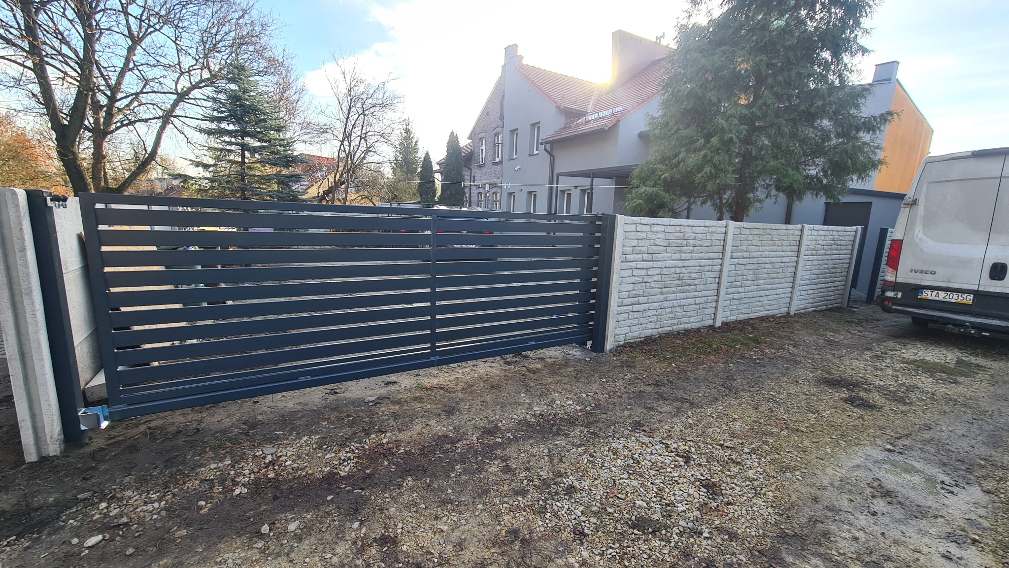 Nowoczesna, przesuwna brama wjazdowa z poziomych profili w kolorze antracytowym, zamontowana między betonowymi słupkami i przylegająca do ogrodzenia z paneli betonowych, na tle domu z czerwonym...