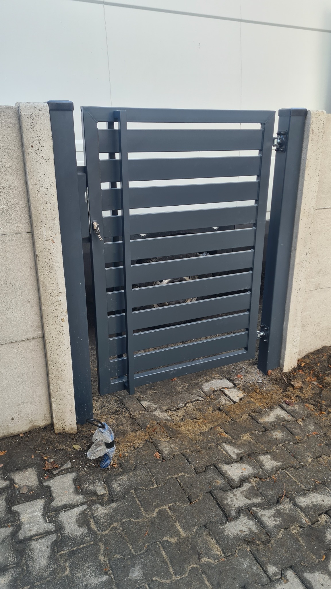 Nowoczesna, szara furtka z poziomych profili, zamontowana między betonowymi słupkami, widoczna na tle białej ściany budynku. Na ziemi, przy słupku, leży zapakowana rura karbowana.