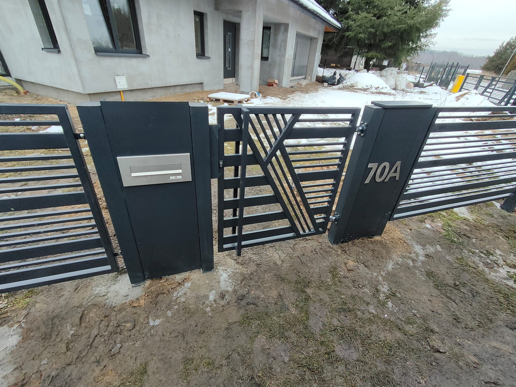 Nowoczesna, czarna brama wjazdowa z geometrycznym wzorem, numerem domu 70A i skrzynką na listy, na tle surowego stanu budynku i resztek śniegu.