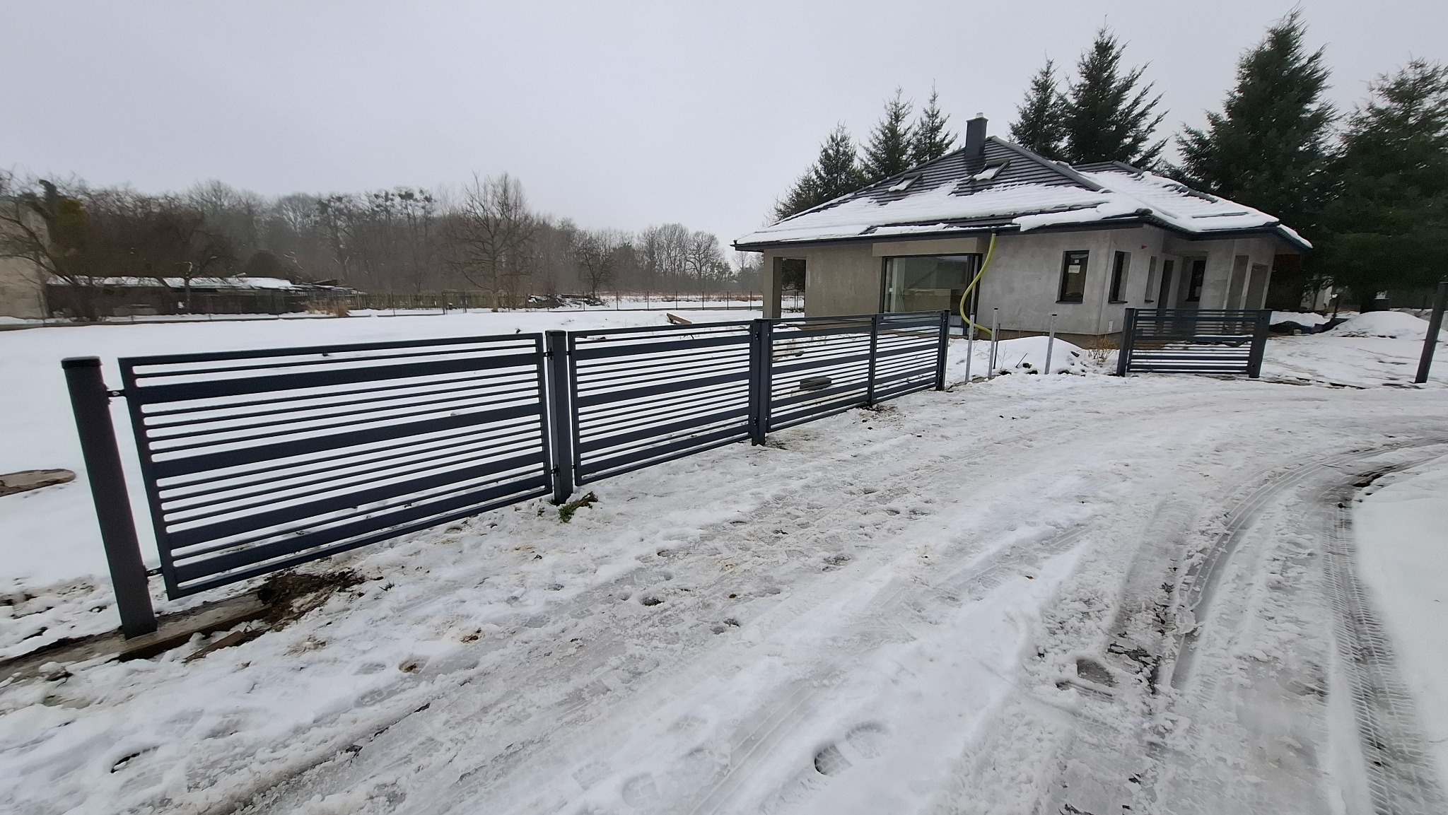 Nowoczesne ogrodzenie panelowe z poziomymi szczeblami w kolorze grafitowym, otoczone śnieżnym krajobrazem przed domem w trakcie budowy.