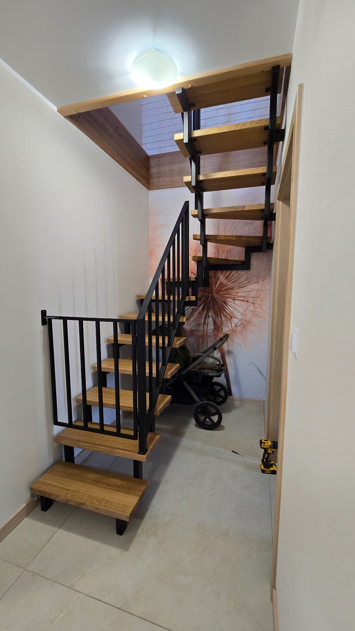 Drewniane schody z czarną metalową konstrukcją i barierką, z zabezpieczeniem dla dzieci na dole. W tle tapeta z motywem dmuchawca i wózek dziecięcy.