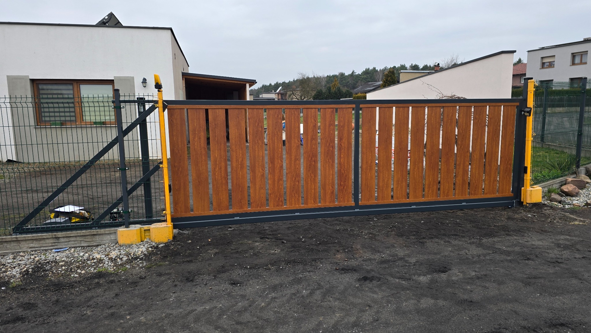 Nowoczesna brama przesuwna z drewnopodobnymi panelami pionowymi i szarą ramą, z żółtymi elementami automatyki, na tle domu z ogrodzeniem panelowym.