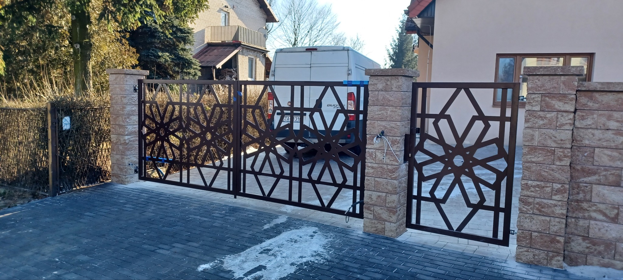 Nowoczesna brama wjazdowa z metalu o geometrycznym wzorze, osadzona na murowanych słupkach. Widoczny automat do bramy i kostka brukowa na podjeździe.
