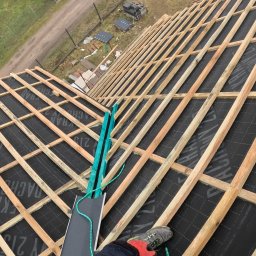 DOMY NA WYMIAR SPÓŁKA Z OGRANICZONĄ ODPOWIEDZIALNOŚCIĄ - Konstrukcja dachu z drewnianych łat i kontrłat pokryta membraną dachową, widok z góry, w tle budynki gospodarcze i zieleń.