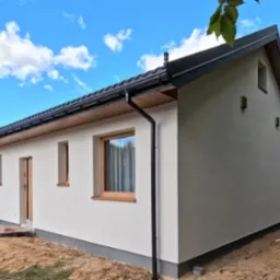 Świeżo otynkowany, parterowy dom jednorodzinny z brązowymi oknami i czarną rynną, widoczny z boku na tle błękitnego nieba z chmurami.