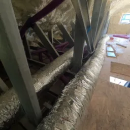 Poddasze z widoczną izolacją natryskową, metalowymi elementami konstrukcyjnymi, fioletowymi rurami wentylacyjnymi i srebrnymi przewodami izolowanymi termicznie, okno dachowe w tle