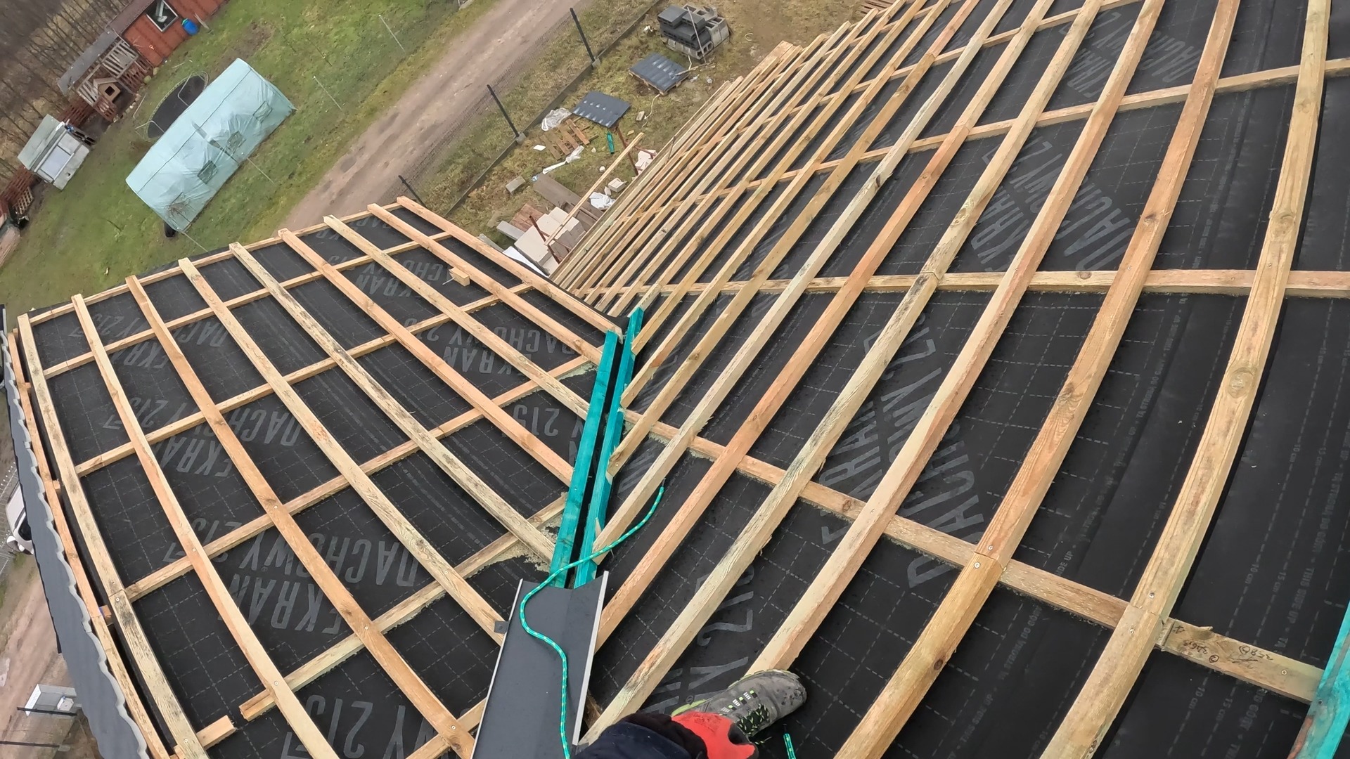 Konstrukcja dachu z drewnianych łat i kontrłat pokryta membraną dachową, widok z góry, w tle budynki gospodarcze i zieleń.