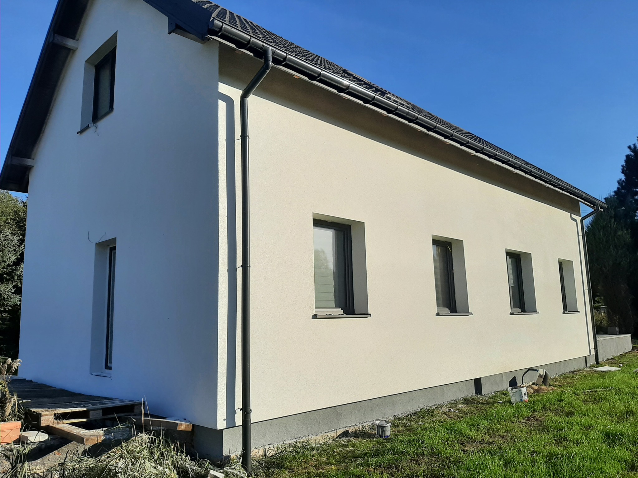 Świeżo otynkowana elewacja domu jednorodzinnego w kolorze kremowym, z widocznymi czarnymi rynnami i ramami okiennymi, na tle błękitnego nieba. Paleta i resztki materiałów budowlanych w pobliżu ściany.