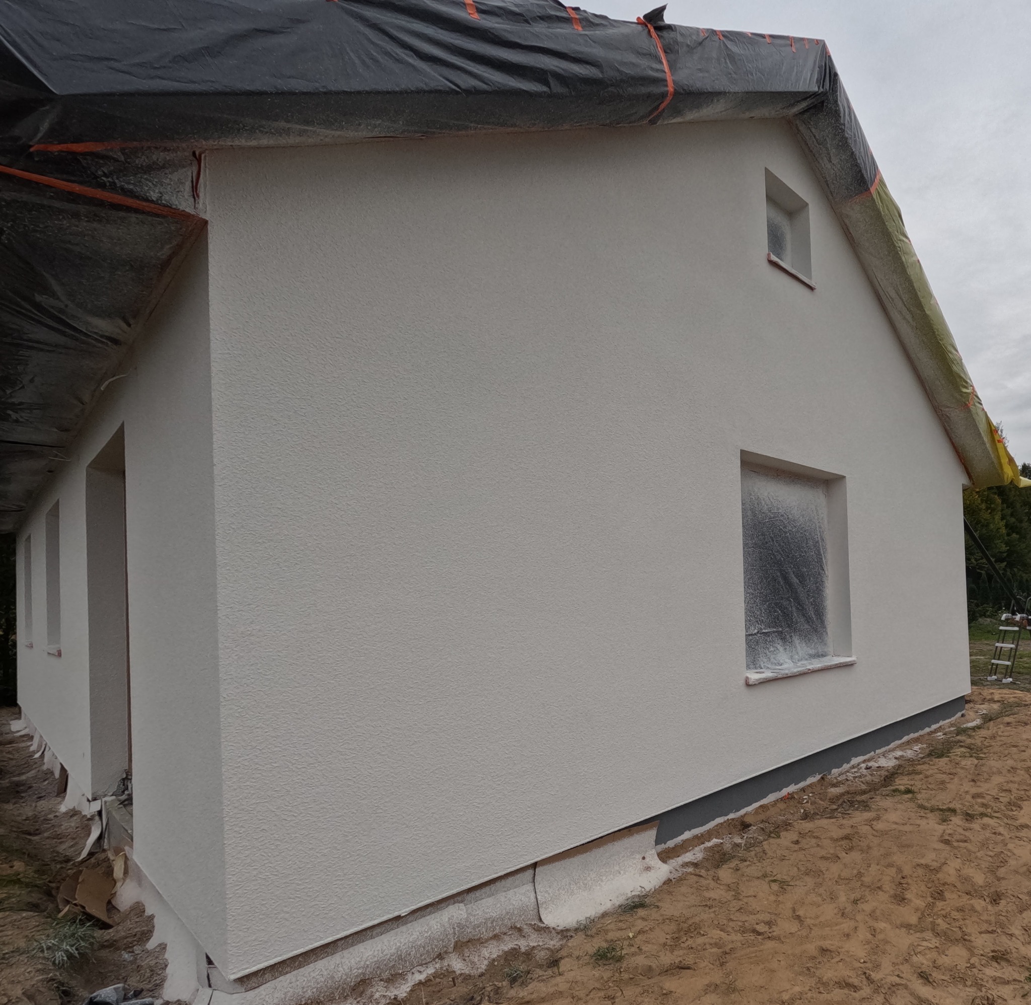 Świeżo otynkowana elewacja domu jednorodzinnego w trakcie prac wykończeniowych, okna zabezpieczone folią, widoczne ślady zaprawy na gruncie, dach przykryty czarną folią ochronną.