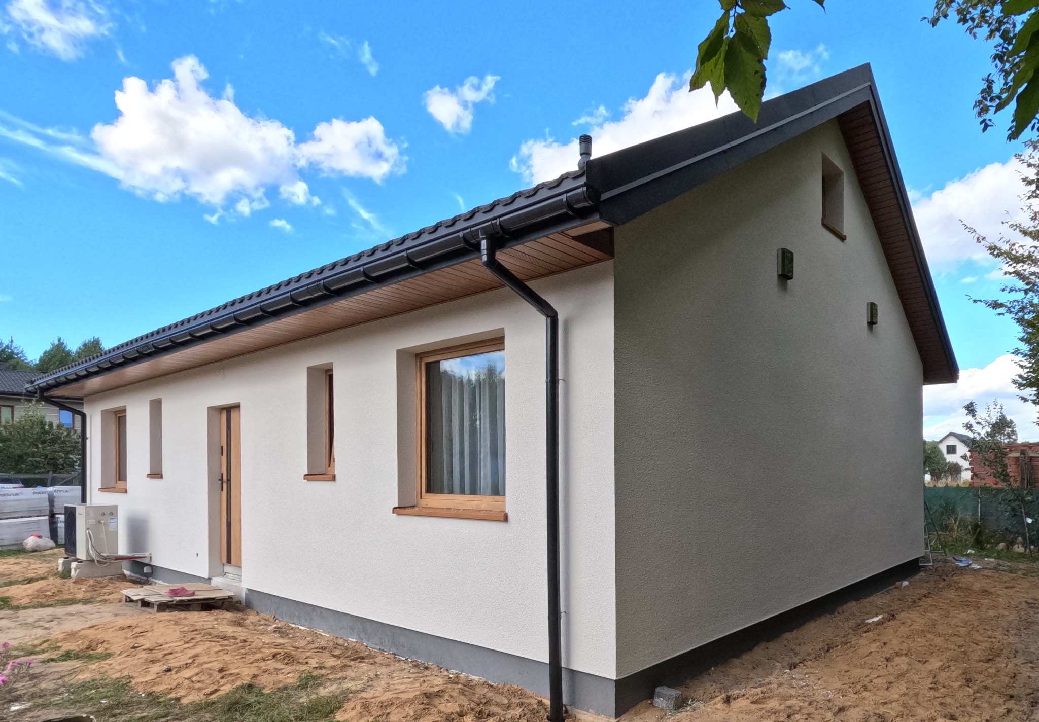 Świeżo otynkowany, parterowy dom jednorodzinny z brązowymi oknami i czarną rynną, widoczny z boku na tle błękitnego nieba z chmurami.