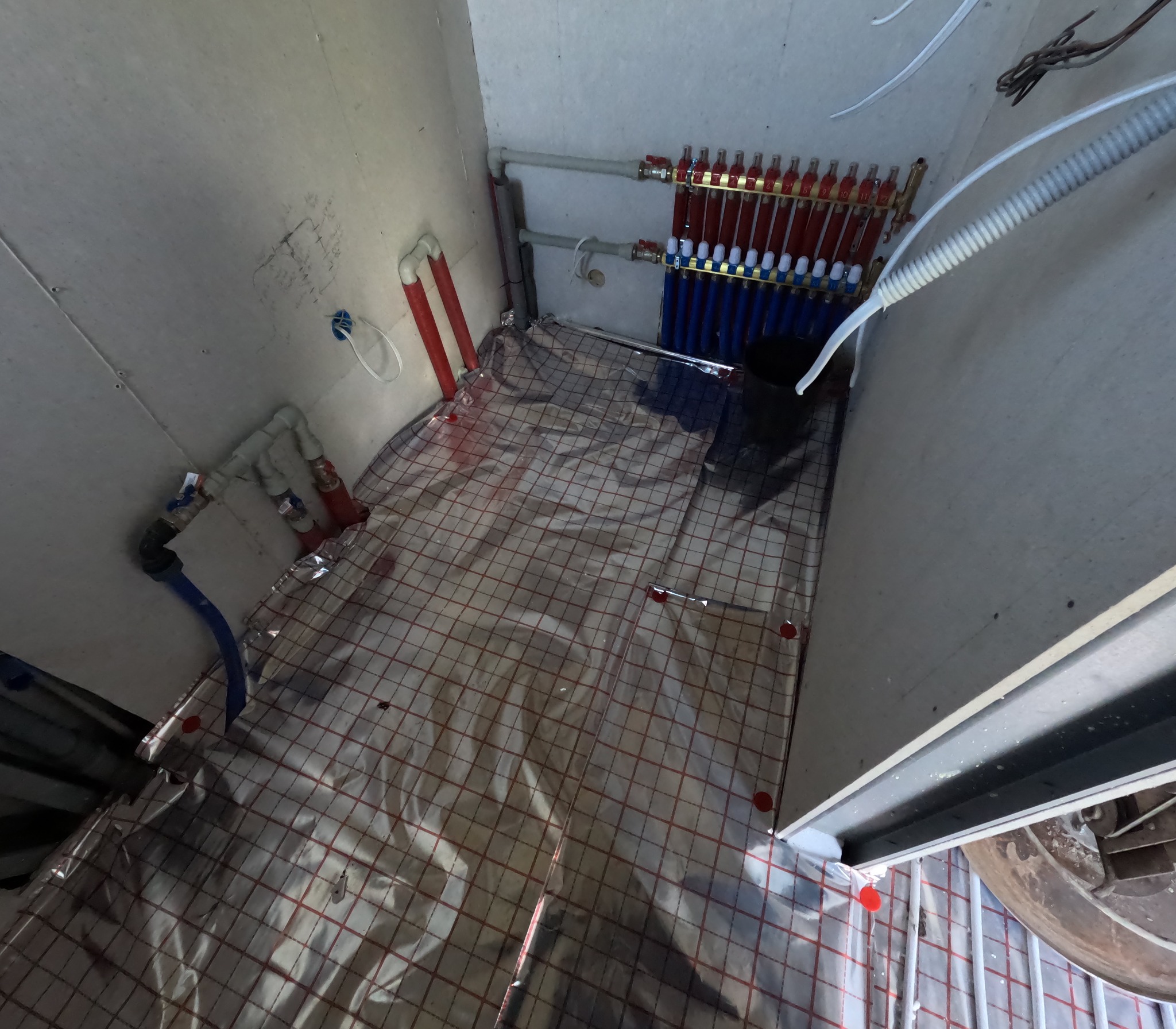 Rozdzielacz instalacji centralnego ogrzewania z rurami PEX ułożonymi na izolacji podłogowej z folią aluminiową i siatką w pomieszczeniu w trakcie montażu.