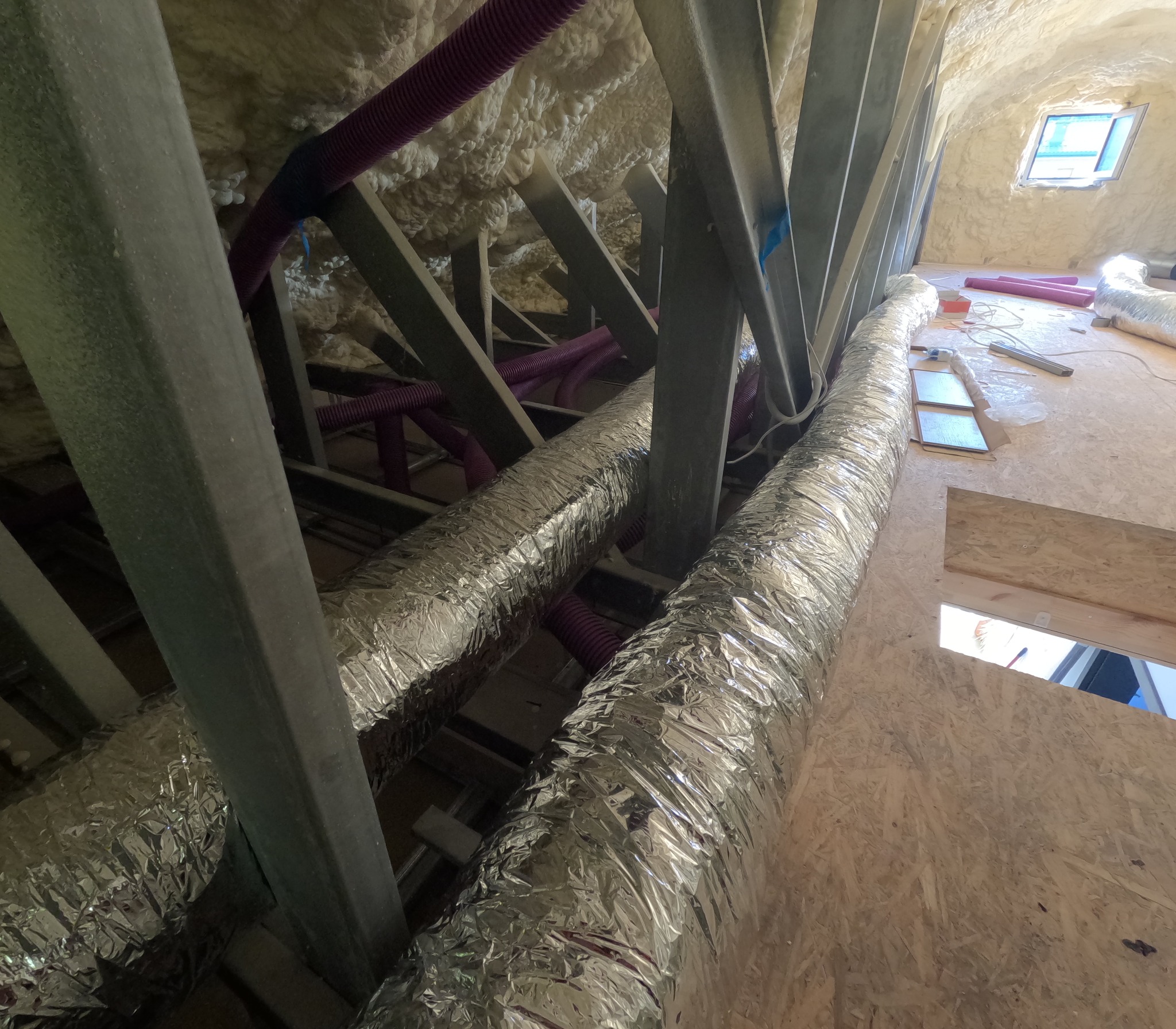 Poddasze z widoczną izolacją natryskową, metalowymi elementami konstrukcyjnymi, fioletowymi rurami wentylacyjnymi i srebrnymi przewodami izolowanymi termicznie, okno dachowe w tle