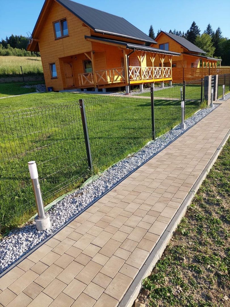 Ułożona kostka brukowa tworząca ścieżkę do drewnianych domków, oddzielona od trawnika ozdobnym kamieniem i oświetlona lampami. Całość otoczona siatką ogrodzeniową.