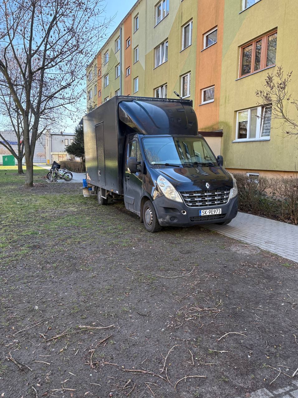Czarny bus Renault Master z wysoką zabudową zaparkowany na trawniku przed blokiem mieszkalnym. Widoczny numer rejestracyjny i fragment chodnika.