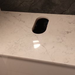 GraniMar - Jasny blat z konglomeratu z otworem na zlew, zamontowany na białej szafce, z ciemnym, kamiennym wykończeniem ściany w tle.