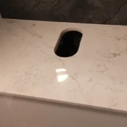 Jasny blat z konglomeratu z otworem na zlew, zamontowany na białej szafce, z ciemnym, kamiennym wykończeniem ściany w tle.