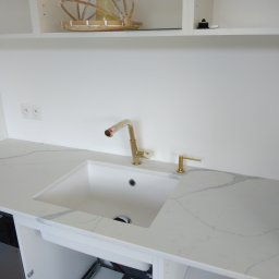 GraniMar - Biały zlew wpuszczony w blat z konglomeratu kwarcowego, złota bateria i dozownik na mydło, minimalistyczna kuchnia z białymi szafkami.