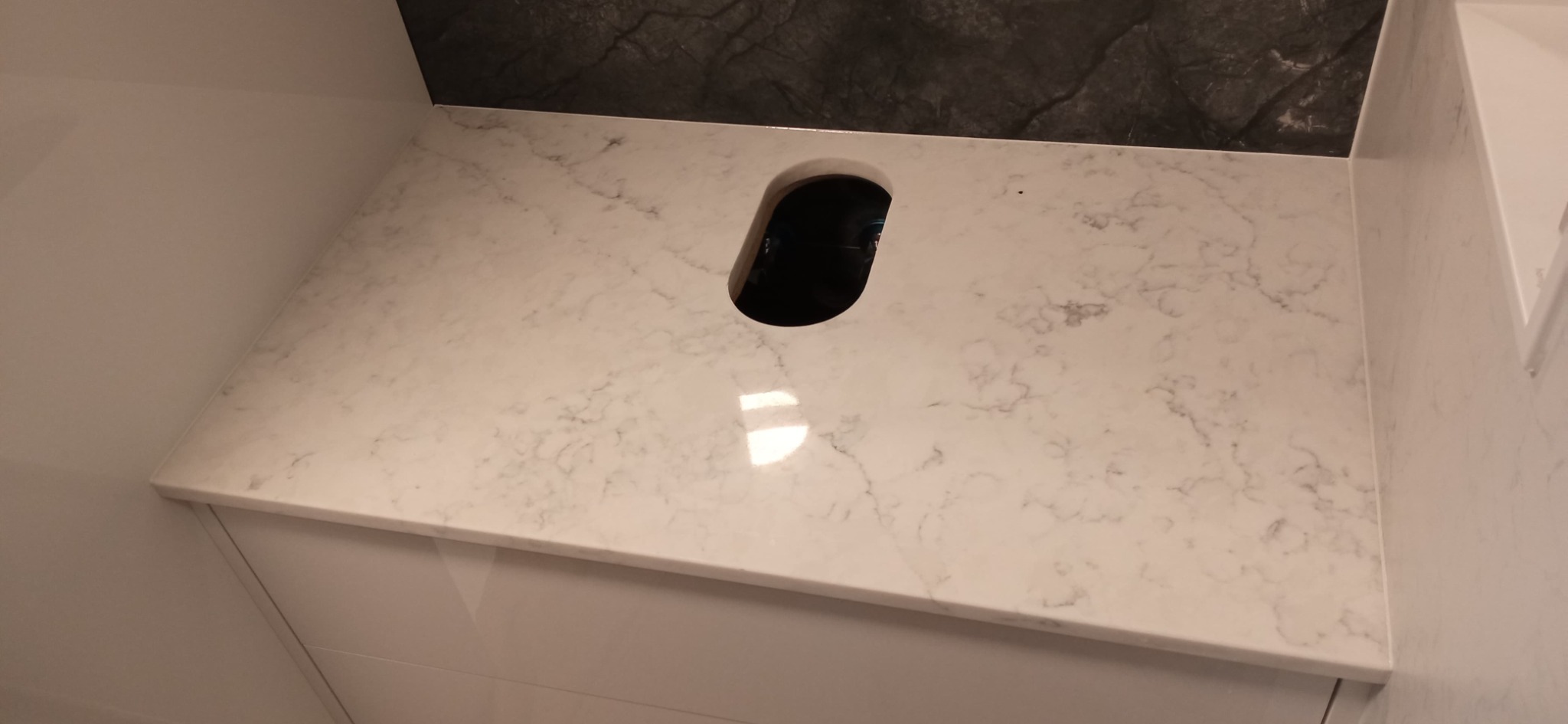 Jasny blat z konglomeratu z otworem na zlew, zamontowany na białej szafce, z ciemnym, kamiennym wykończeniem ściany w tle.