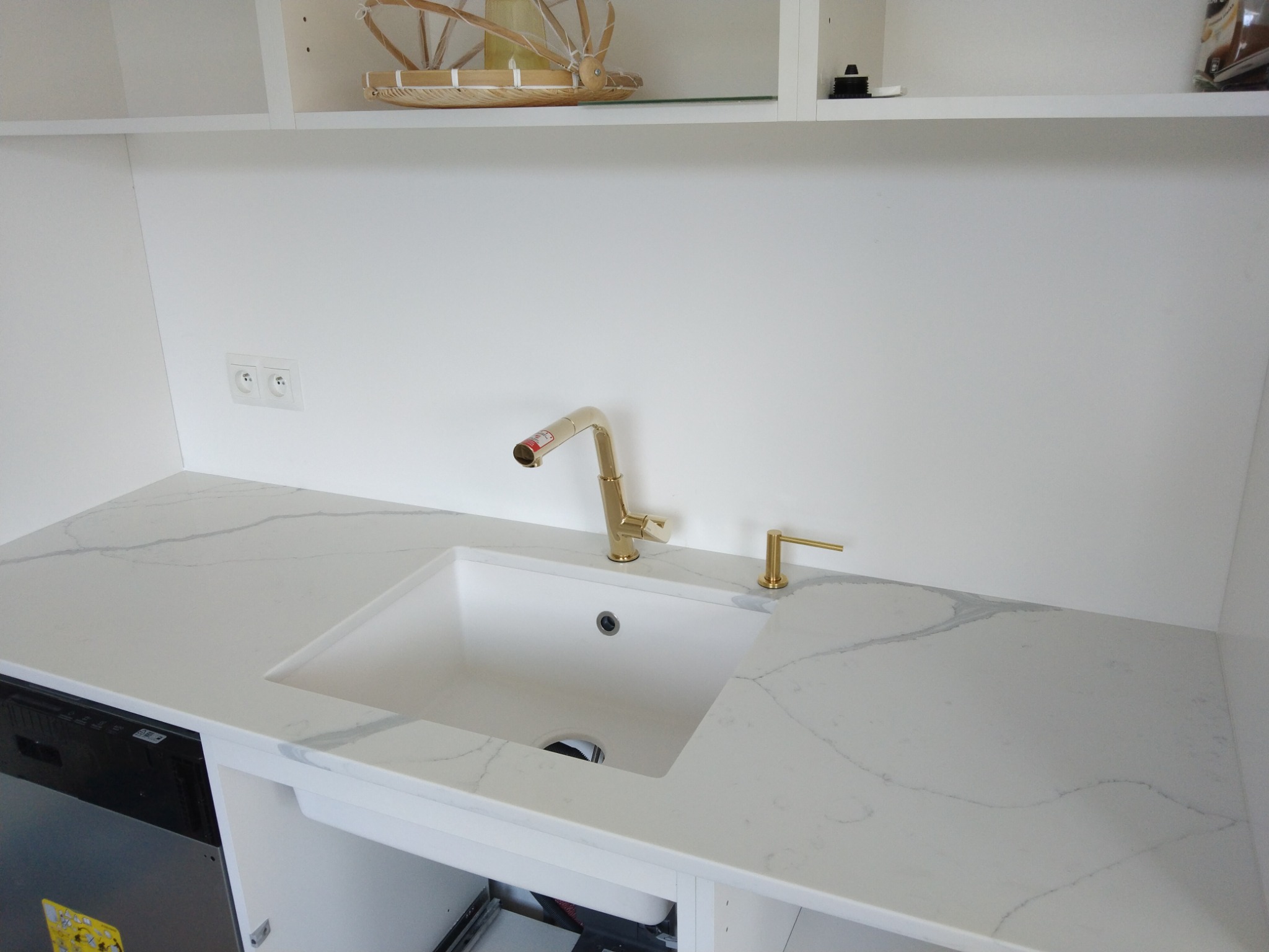 Biały zlew wpuszczony w blat z konglomeratu kwarcowego, złota bateria i dozownik na mydło, minimalistyczna kuchnia z białymi szafkami.