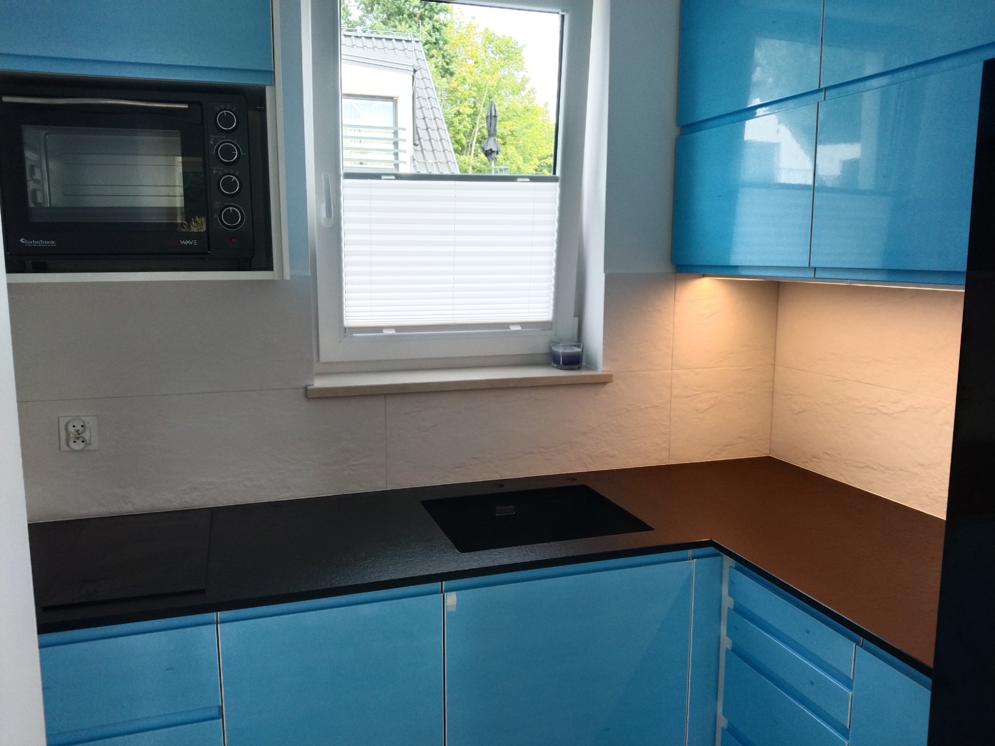 Czarny kamienny blat kuchenny z wbudowanym zlewozmywakiem, w nowoczesnej kuchni z błękitnymi szafkami i białymi płytkami ściennymi, oświetlony ciepłym światłem spod szafek.