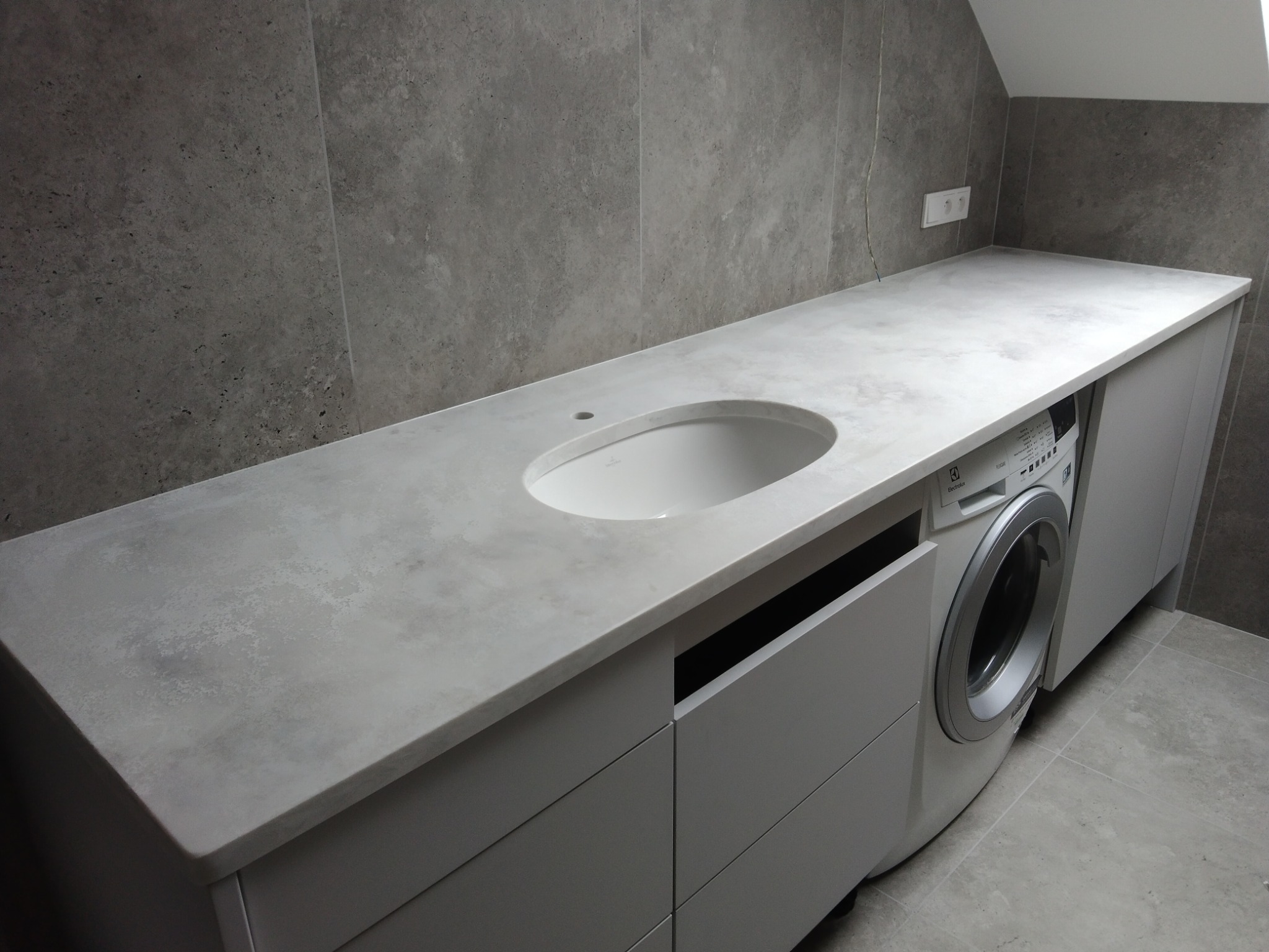 Szary blat z umywalką wpuszczaną w minimalistycznej łazience z pralką pod blatem.