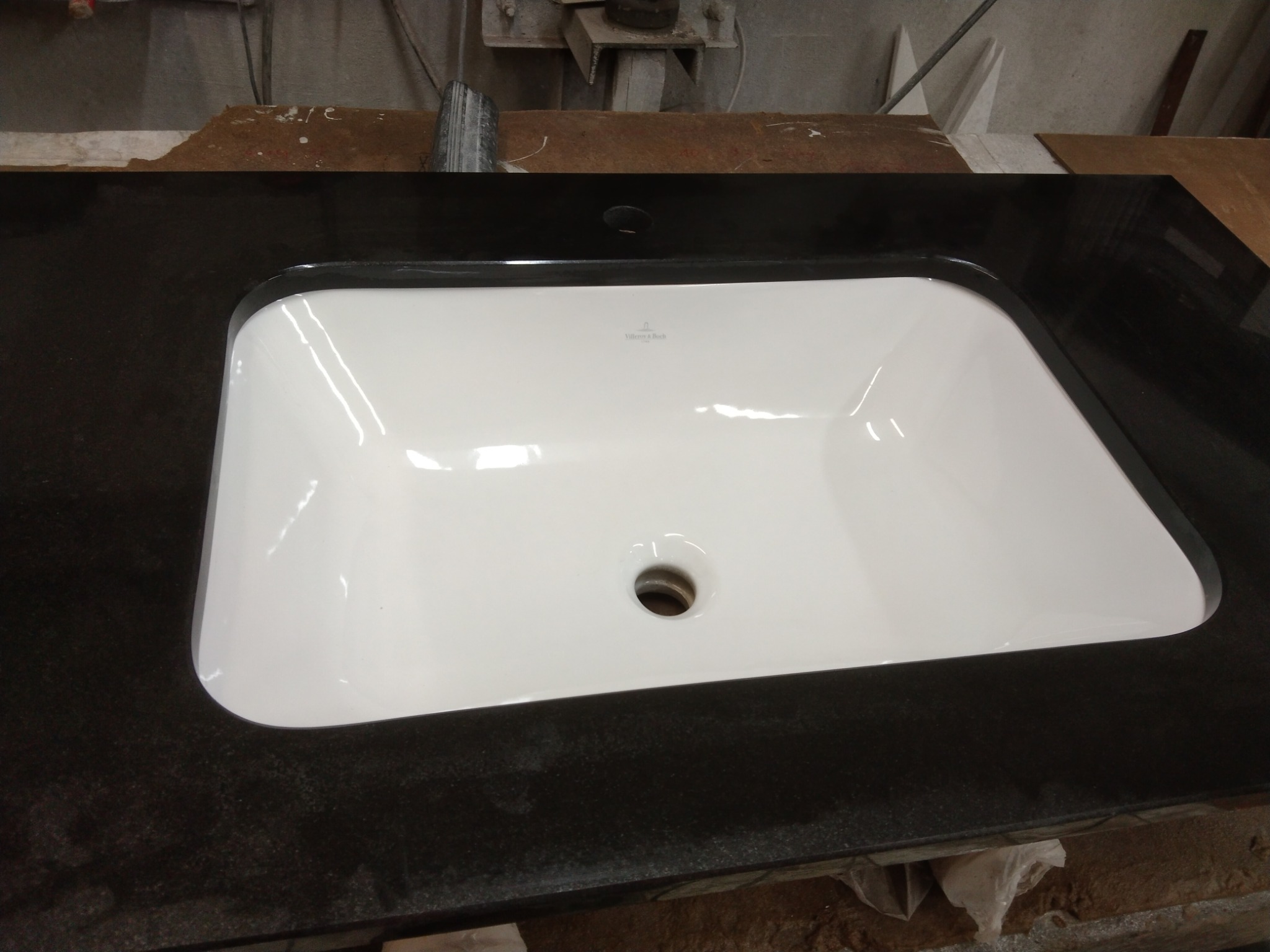 Biała umywalka Villeroy & Boch wpuszczona w czarny, polerowany blat z kamienia, widoczne otwory na baterię, w tle warsztat kamieniarski.
