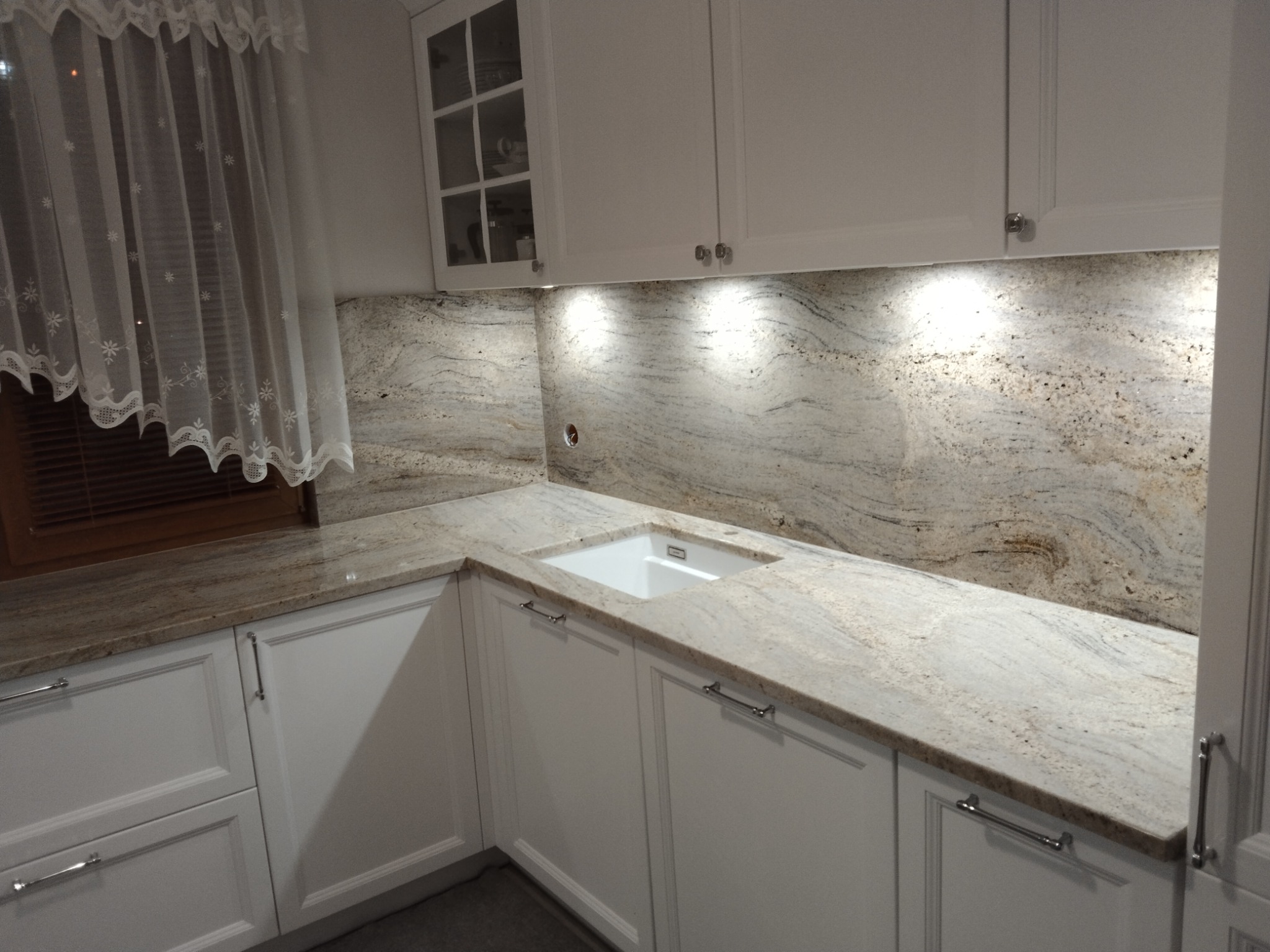 Jasny, wzorzysty blat kuchenny z kamienia naturalnego z wbudowanym zlewem, podświetlony punktowymi lampkami podszafkowymi, na tle białych szafek kuchennych i firanki w oknie.