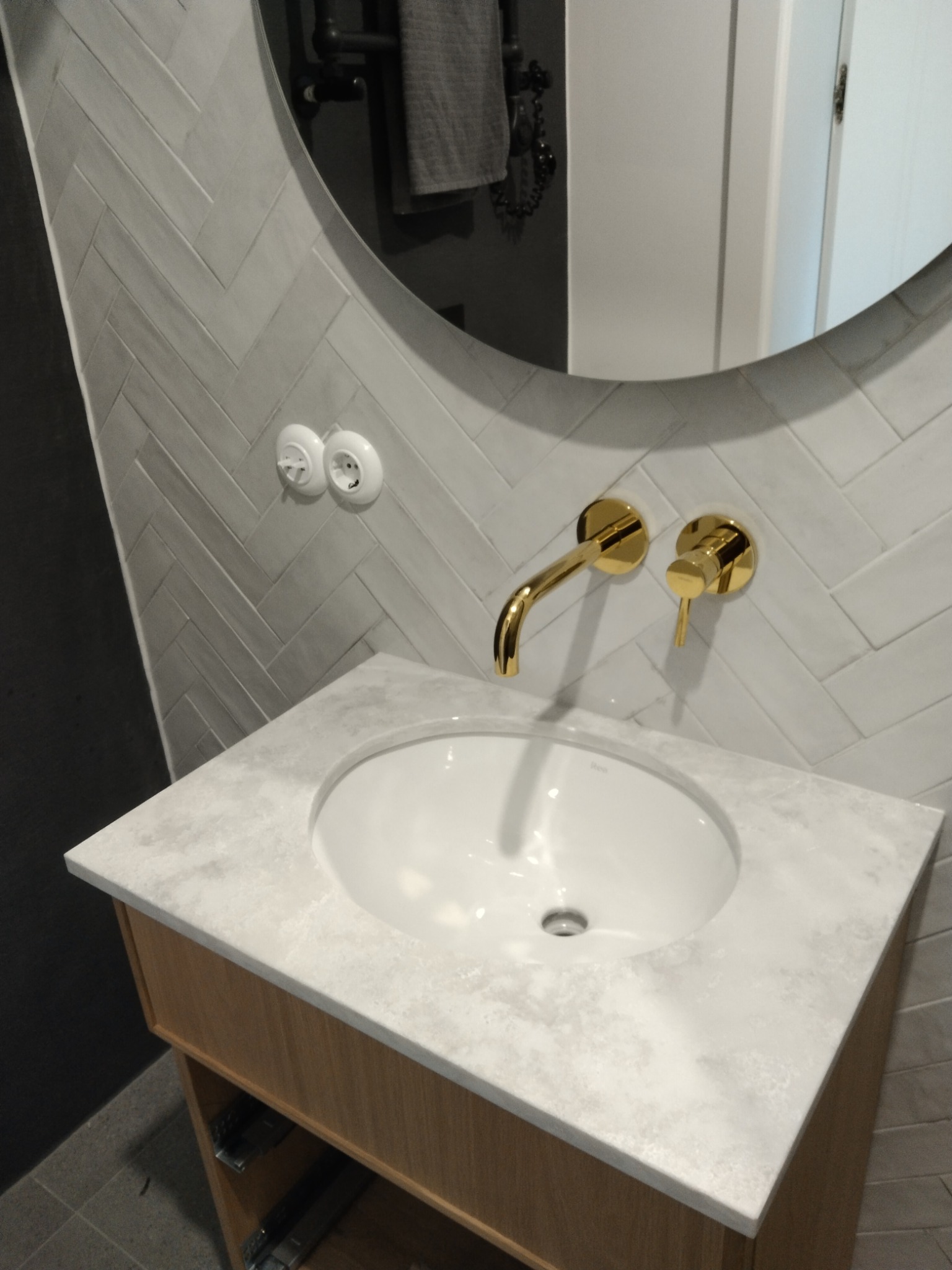 Łazienka z umywalką nablatową na drewnianej szafce, złota armatura na tle ściany z szarej cegły ułożonej w jodełkę.