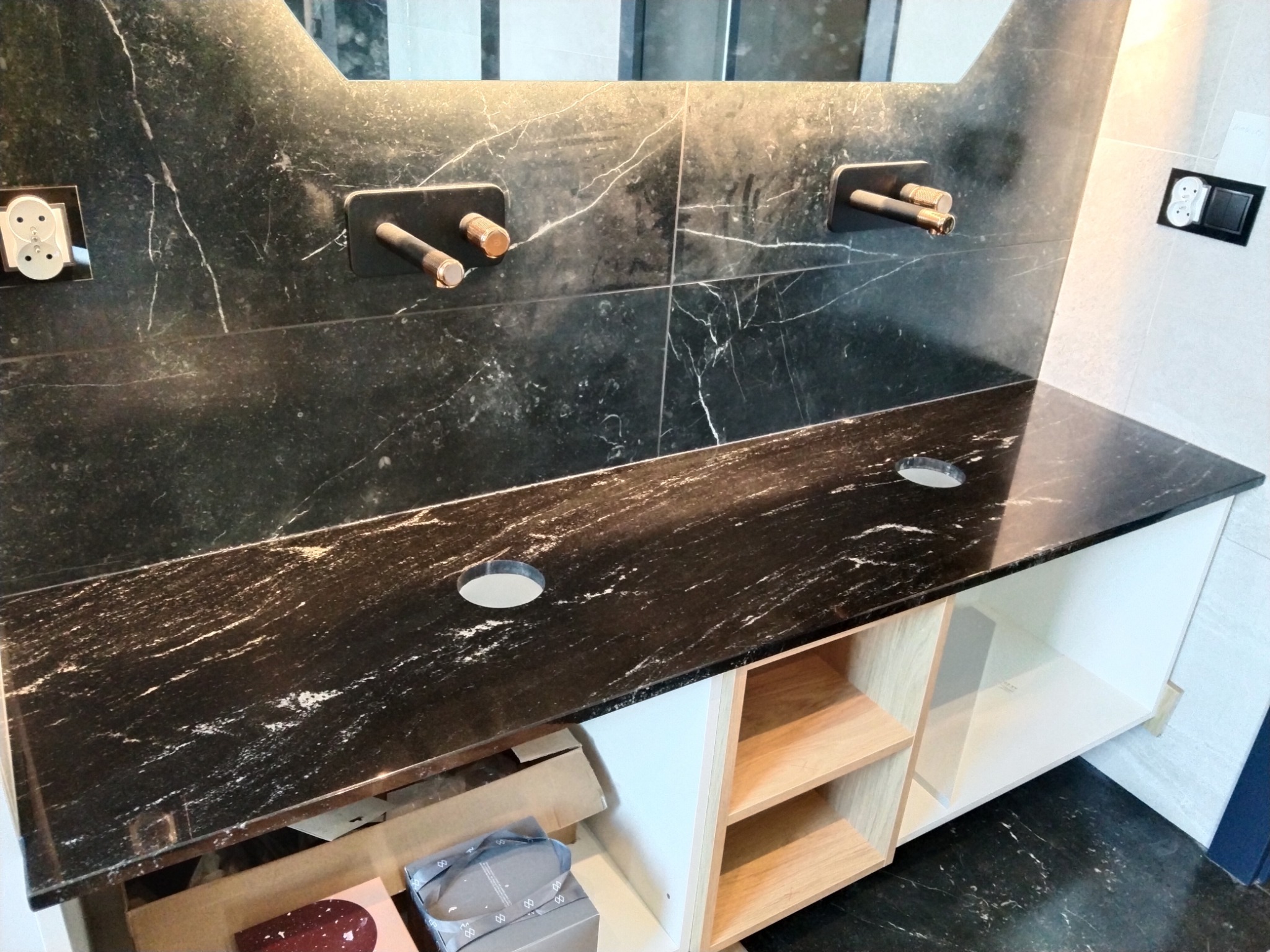 Czarny blat z marmurowym wzorem z wyciętymi otworami na umywalki i baterie, zamontowany na szafce z jasnego drewna z widocznymi elementami instalacji wodnej.