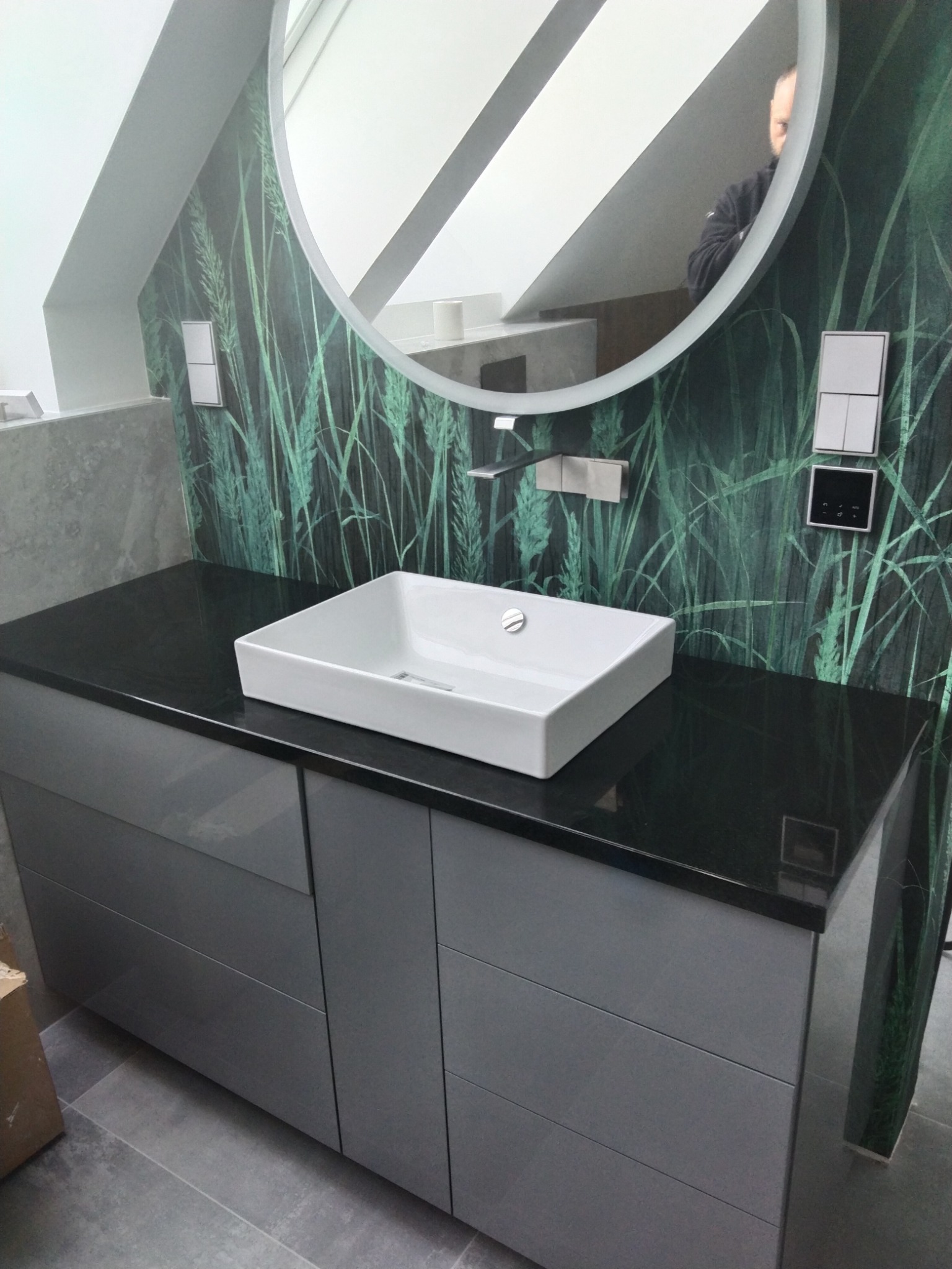 Łazienka z czarnym blatem, białą, kwadratową umywalką, szarą szafką pod umywalką i okrągłym lustrem z podświetleniem LED na ścianie z motywem trawy.