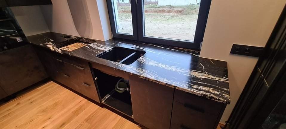 Czarny blat kamienny z białymi żyłkami, zamontowany w kuchni, z widocznym zlewem i oknem w tle, drewniana podłoga.