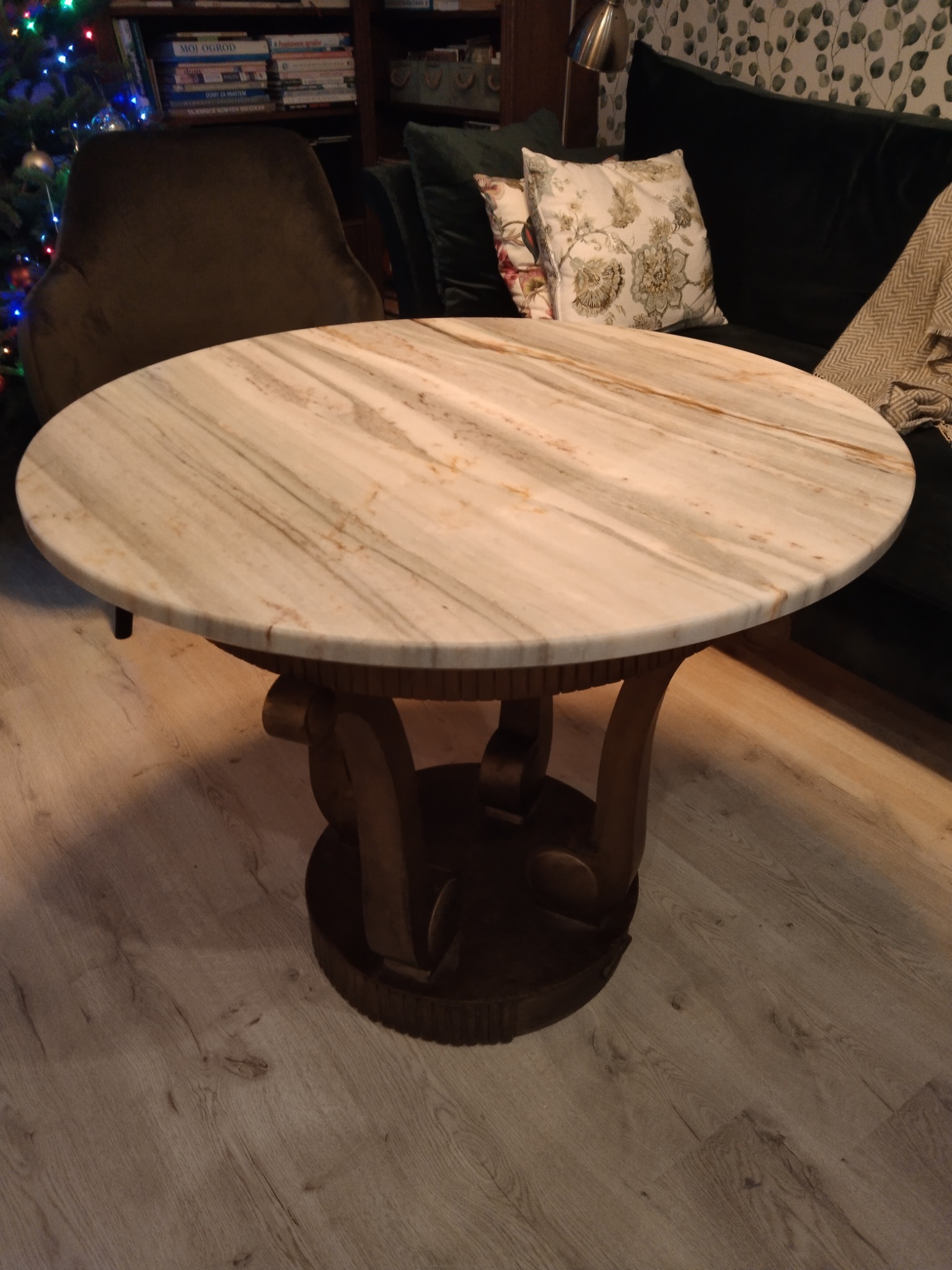Okrągły stolik kawowy z blatem z jasnego marmuru z wyraźnym żyłkowaniem, na ozdobnej, drewnianej podstawie, w tle sofa z poduszkami i choinka.