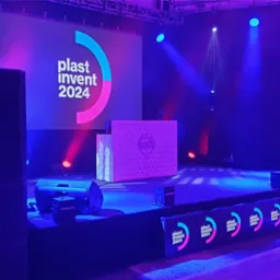 Scena oświetlona niebieskim i różowym światłem, z kolumnami głośnikowymi po bokach, podświetlanym DJ-skim stanowiskiem na środku i ekranem z logo 'plast invent 2024' w tle oraz logotypem 'TEAM...