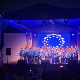 SOUNDTEAM JUSTYNA MOLENDA - Chór dziecięcy na scenie podczas występu, widoczne oświetlenie sceniczne i telebim z motywem flag europejskich, logo firmy nagłośnieniowej Team Sound w lewym górnym rogu.