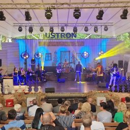 SOUNDTEAM JUSTYNA MOLENDA - Scena plenerowa z zespołem muzycznym podczas występu, publiczność siedząca na ławkach, oświetlenie sceniczne i dekoracje, logo Team Sound w prawym górnym rogu.