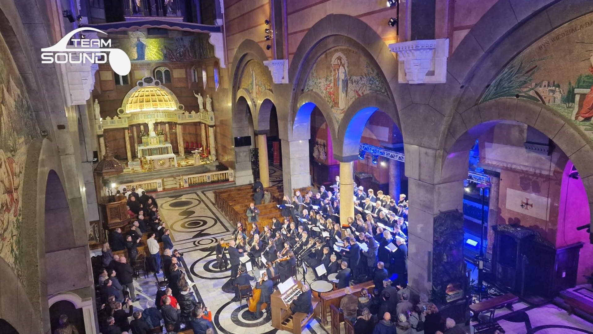 Chór i orkiestra w kościele z nagłośnieniem i oświetleniem, widok z góry na zespół muzyczny i publiczność.