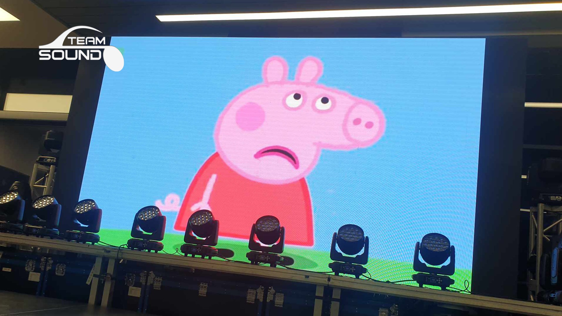 Duży ekran LED wyświetla postać z kreskówki Peppa Pig z wyrazem smutku, poniżej rząd czarnych reflektorów scenicznych na metalowej konstrukcji.