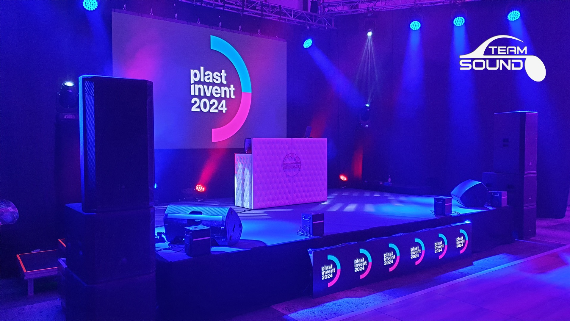 Scena oświetlona niebieskim i różowym światłem, z kolumnami głośnikowymi po bokach, podświetlanym DJ-skim stanowiskiem na środku i ekranem z logo 'plast invent 2024' w tle oraz logotypem 'TEAM...