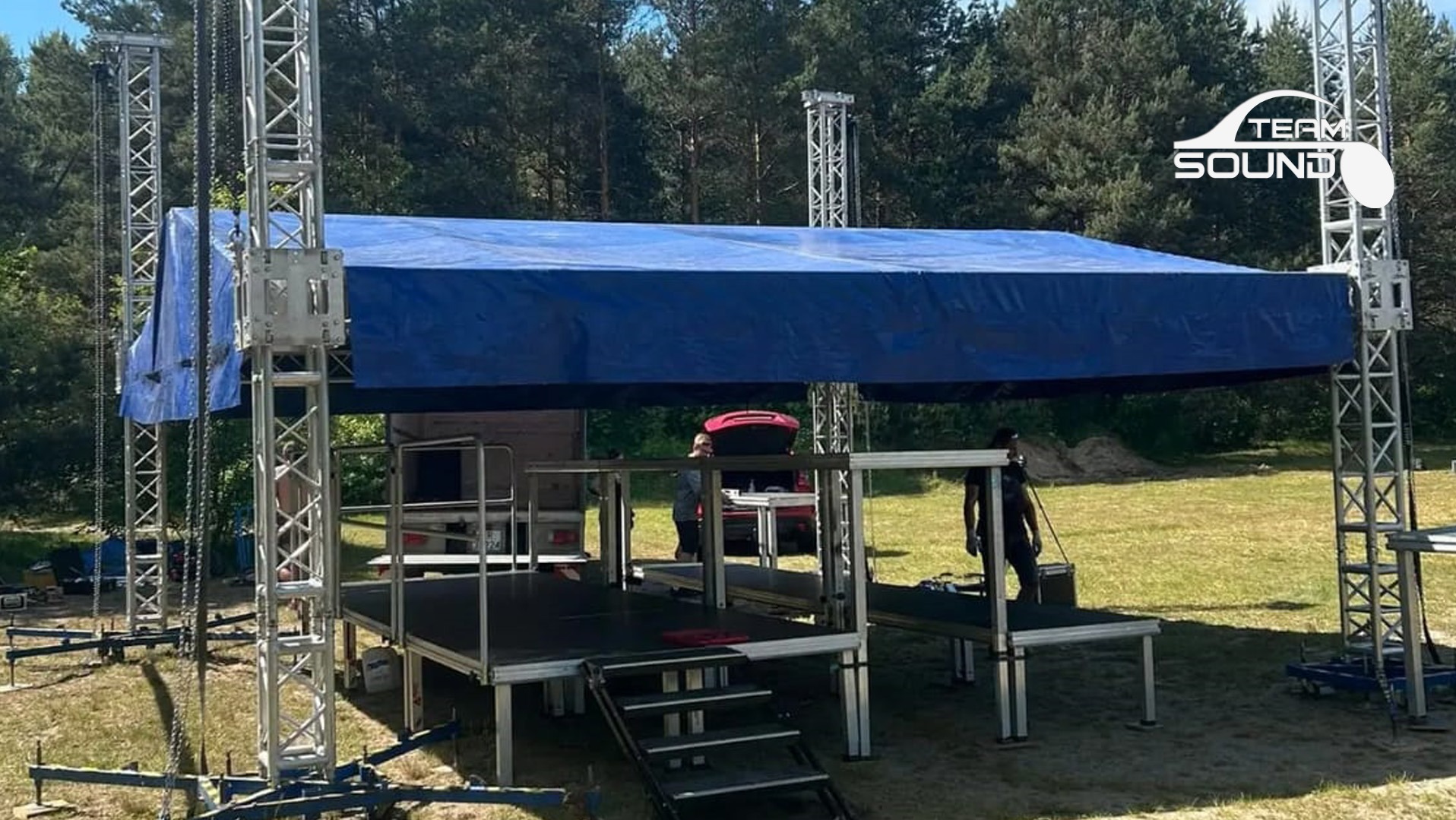 Montaż sceny plenerowej z zadaszeniem z niebieskiej plandeki, widoczne stalowe konstrukcje nośne i podest, w tle las i częściowo widoczny samochód z otwartym bagażnikiem.
