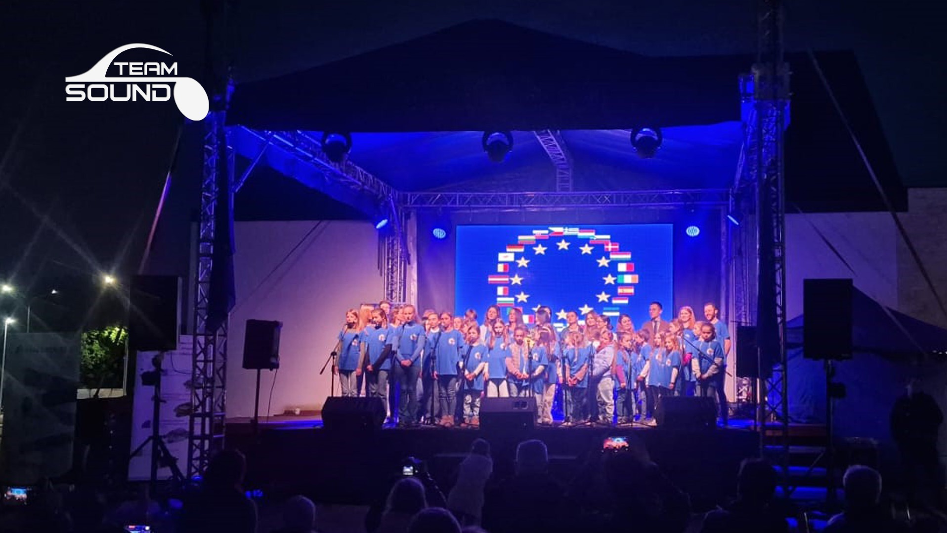 Chór dziecięcy na scenie podczas występu, widoczne oświetlenie sceniczne i telebim z motywem flag europejskich, logo firmy nagłośnieniowej Team Sound w lewym górnym rogu.