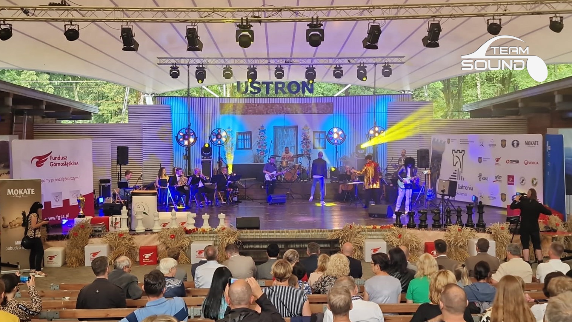 Scena plenerowa z zespołem muzycznym podczas występu, publiczność siedząca na ławkach, oświetlenie sceniczne i dekoracje, logo Team Sound w prawym górnym rogu.