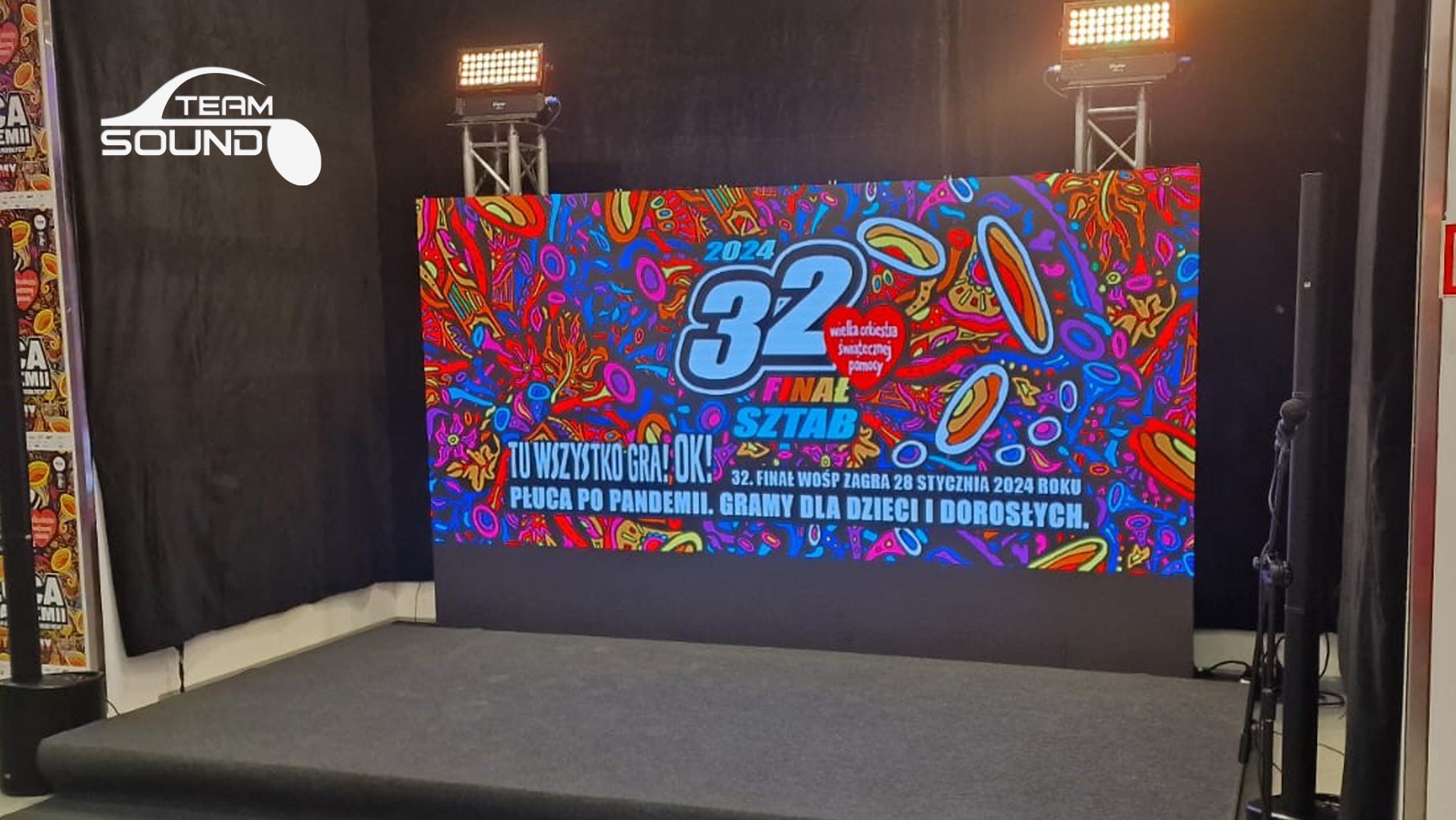Scena z podświetlonym ekranem LED prezentującym logo i hasła 32. Finału WOŚP w 2024 roku, oświetlenie sceniczne na statywach, czarne tło i nagłośnienie boczne.