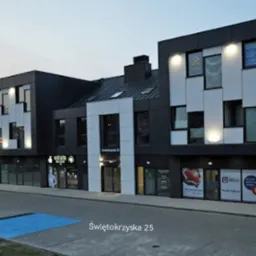 Nowoczesny budynek biurowy z lokalami usługowymi na parterze, widoczne szyldy firm i oświetlenie zewnętrzne.