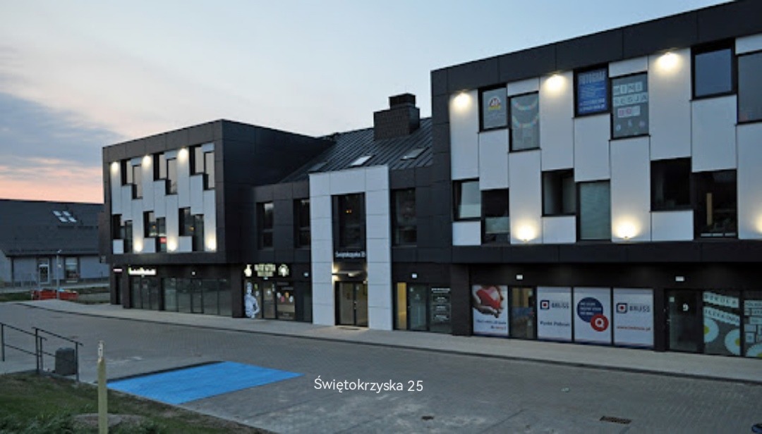 Nowoczesny budynek biurowy z lokalami usługowymi na parterze, widoczne szyldy firm i oświetlenie zewnętrzne.